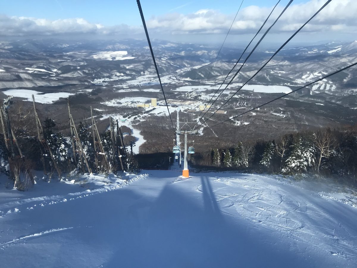 從安比纜車上往下看:纜線與一座橘色底座的支柱沿著雪坡往下延伸,坡道兩側是掛霜的樹林,遠處山谷裡看得到 Crowne Plaza 一帶的黃色建築,再遠是一層層覆雪的群山