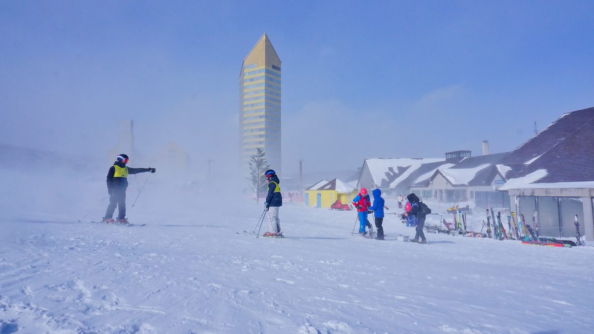 風雪中的山腳緩坡,一位穿黃色背心、戴安全帽與護目鏡的大人伸手指方向,對面一個同樣穿黃色背心的小孩拄著雪杖站在雪板上,右邊幾個孩子在休息區旁,背景是 Crowne Plaza 的黃色高樓