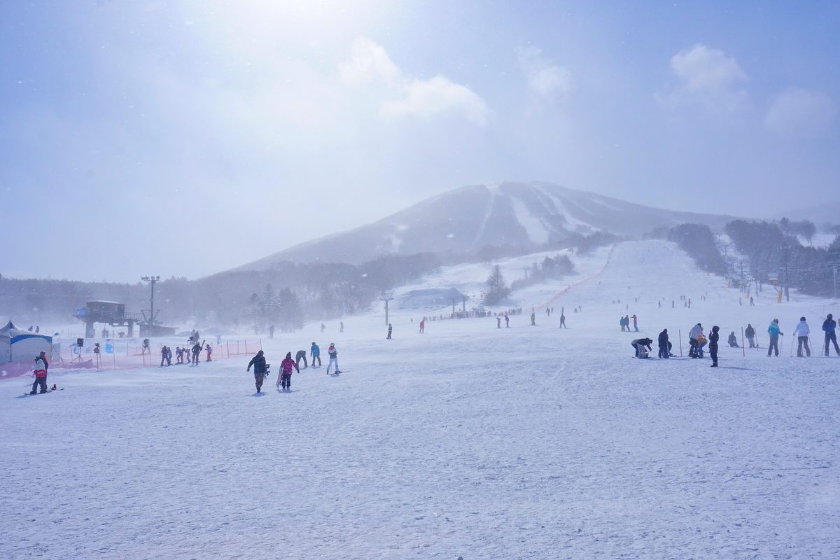 安比高原滑雪場山腳的寬闊緩坡,正前方是覆雪的前森山,山坡上幾條雪道被風雪遮得朦朧;坡底散著幾十位穿雪衣、戴護目鏡的滑雪者,左側有吊椅站與藍色帳篷