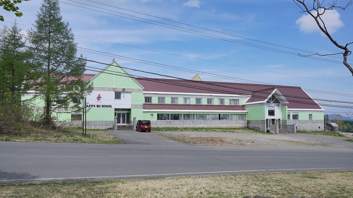 路旁一棟淺綠色外牆、暗紅色屋頂的兩層長形建築,左側山牆下寫著 APPI SKI SCHOOL,門前停著一輛紅色小車,周圍沒有雪