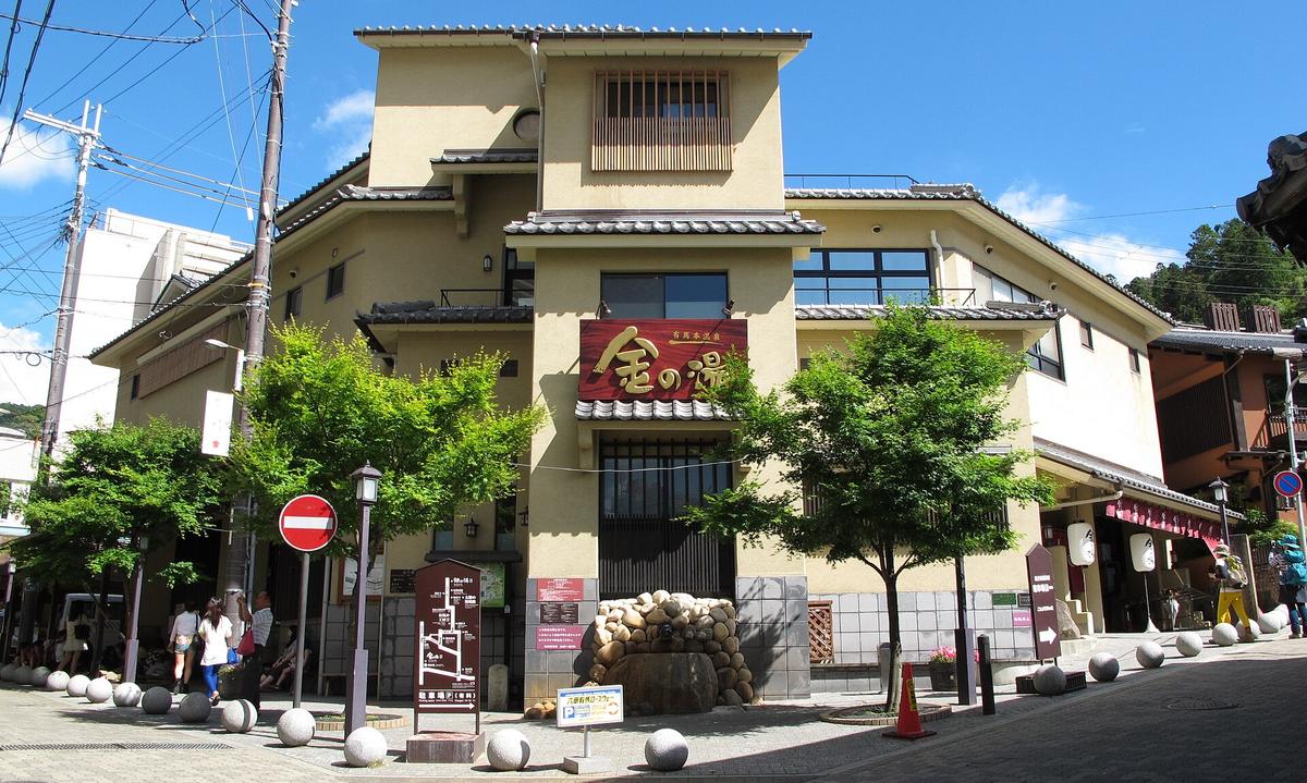 有馬溫泉「金の湯」外觀：土黃色外牆的三層和風建築，正面掛著紅底金字的「有馬本温泉 金の湯」招牌，門前有一座卵石堆成的造景與成排的石球車阻，前方路口立著一塊寫「六甲有馬ロープウェー のりば」的指示牌，左側樹蔭下有幾位遊客