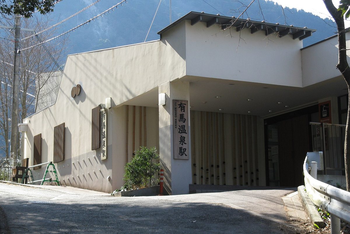 六甲有馬纜車有馬溫泉站的站房：米白色建築，入口柱子上掛著直式木牌「有馬温泉駅 ARIMA ONSEN STN.」，門口右側有一塊橘色的纜車圖示牌，前方是一段上坡的柏油路，左邊的樹落了葉，背後是山