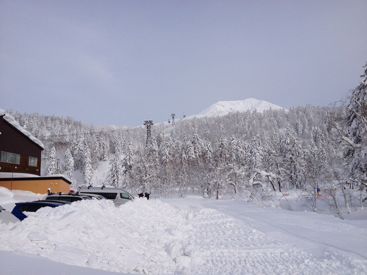冬天的旭岳纜車山麓一帶,停車場的車子半埋在剷起的雪堆後面,背後是積滿雪的針葉林,纜車鐵塔與鋼索往山上延伸,遠處線上掛著一節紅色車廂