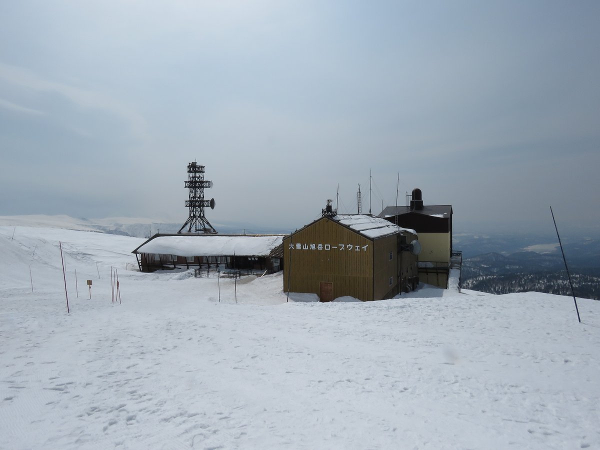 積雪中的旭岳纜車姿見站,土黃色建物側面寫著「大雪山旭岳ロープウェイ」,左邊是屋頂積雪的長形站房與一座通訊鐵塔,右後方是遠處的山谷與平地