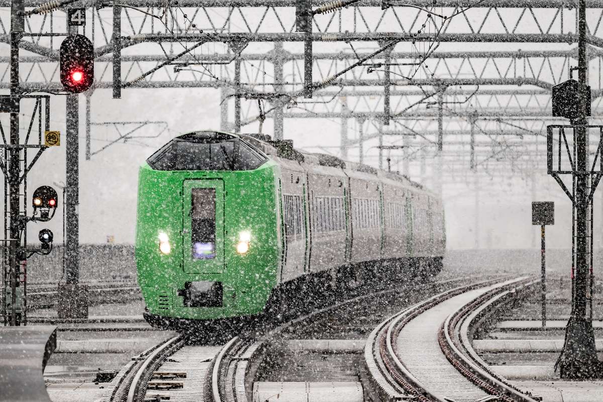 大雪中駛進旭川站的 JR 北海道 789 系特急ライラック,車頭是亮綠色,後面接銀色車廂,上方是整片電車線鐵架與紅色號誌