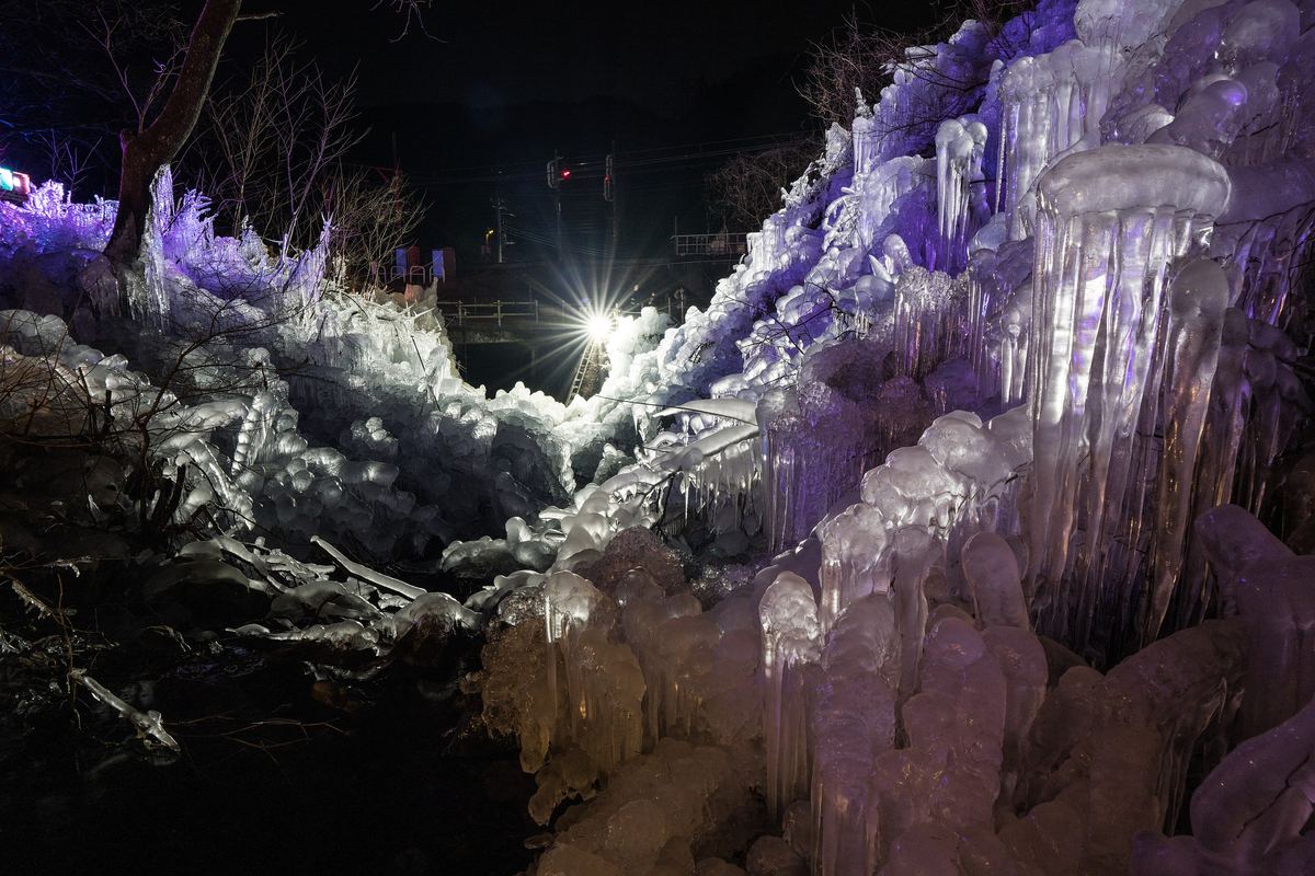 2023 年 1 月蘆久保冰柱夜間點燈的近景:右側一根根垂下的粗冰柱打著白光與淡紫光,冰層沿著山坡往後延伸,遠處一盞強光燈,後方看得到鐵路的紅色號誌