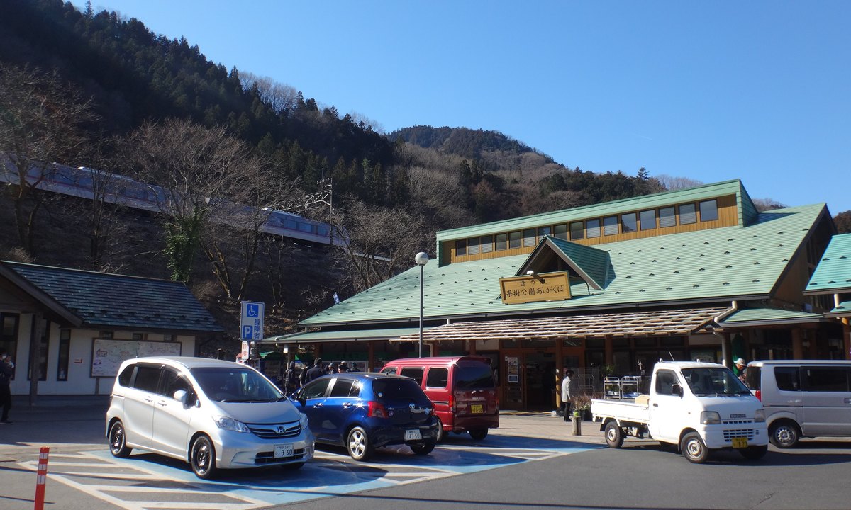 2017 年 2 月晴天的道の駅 果樹公園あしがくぼ:綠色屋頂的木造建築掛著「道の駅 果樹公園あしがくぼ」木招牌,前面是停車場,左後方山坡上的鐵路有一列列車通過