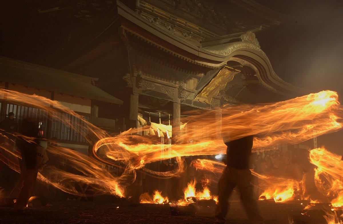 夜裡的阿蘇神社火振り神事：人甩著點燃的茅草束，長時間曝光拉出一圈圈橘色火痕，地上散著燃燒的火堆，背景是阿蘇神社的樓門