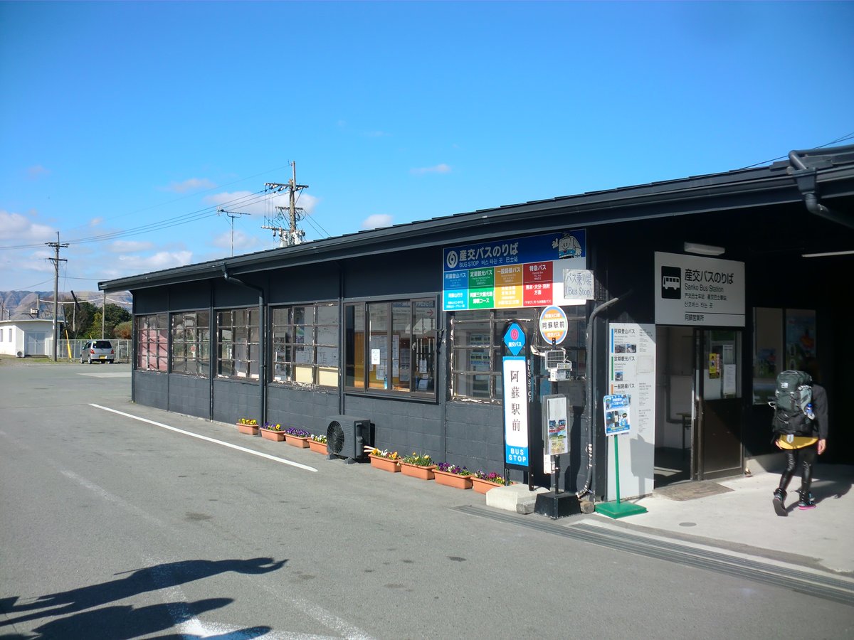 JR 阿蘇駅旁的產交巴士站：深灰色平房，屋簷上的看板寫著「産交バスのりば」並分色列出阿蘇登山バス、定期観光バス、一般路線バス、特急バス，門口立著「阿蘇駅前」站牌，右邊入口掛著「産交バスのりば Sanko Bus Station」