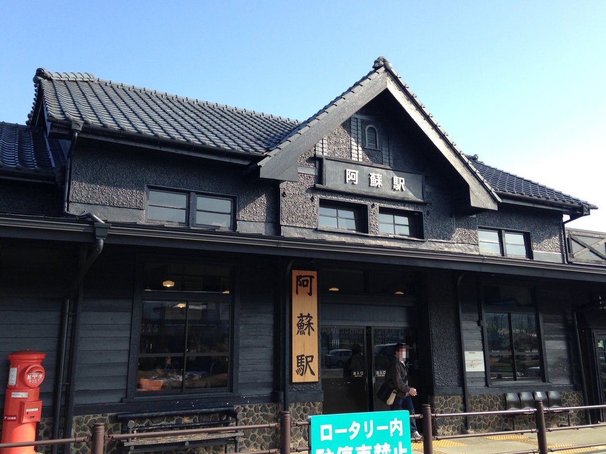 JR 阿蘇駅的站房：黑色木造外牆、瓦屋頂，山牆上掛著「阿蘇駅」站名，入口旁直立的木牌也寫著阿蘇駅，左邊是一個紅色圓筒郵筒，晴天