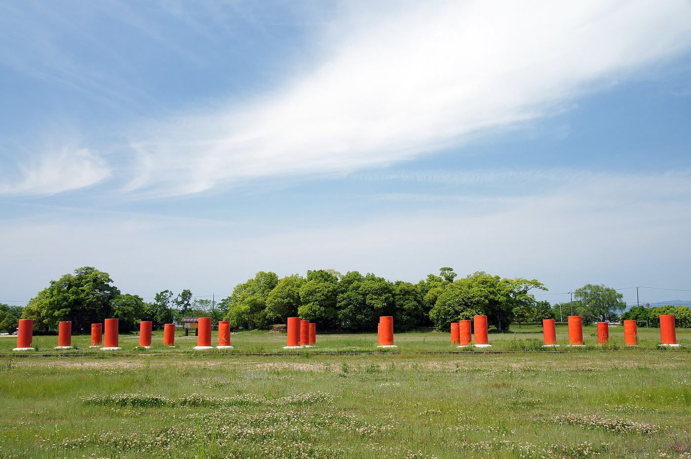The Fujiwara Palace Site: a line of orange-red cylindrical column stumps on square white bases running across a wide grass field, each stump no higher than an adult waist, a row of trees and utility poles behind them and low hills beyond, with sky filling the upper half of the frame
