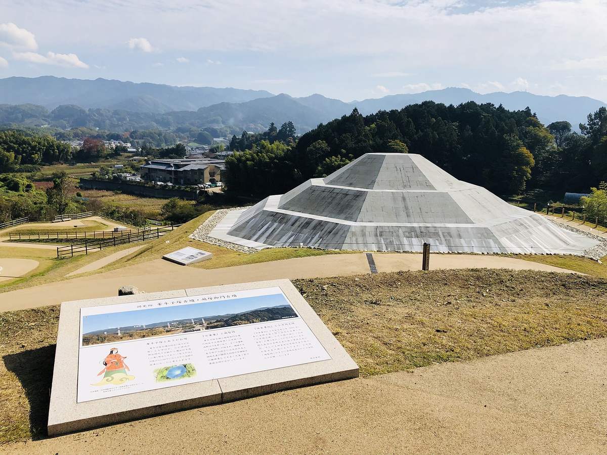 The reconstructed Kengoshizuka Kofun: pale grey cut stone facing laid course by course over a stepped mound rising to a flat top, gravel and sand paths with wooden fencing around it, an interpretive panel set into the ground in the foreground, and a hamlet, fields and layered mountains behind
