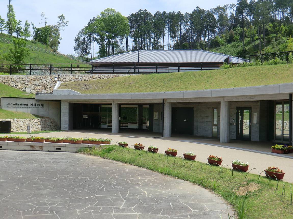 Exterior of the Kitora mural hall Shijin no Yakata: a board-formed concrete building set into a grassed slope so that its roof continues the hillside, the building name carved into the left-hand wall in Japanese and English, a curved flagstone forecourt lined with planters, and a small grey-tiled structure on the slope above