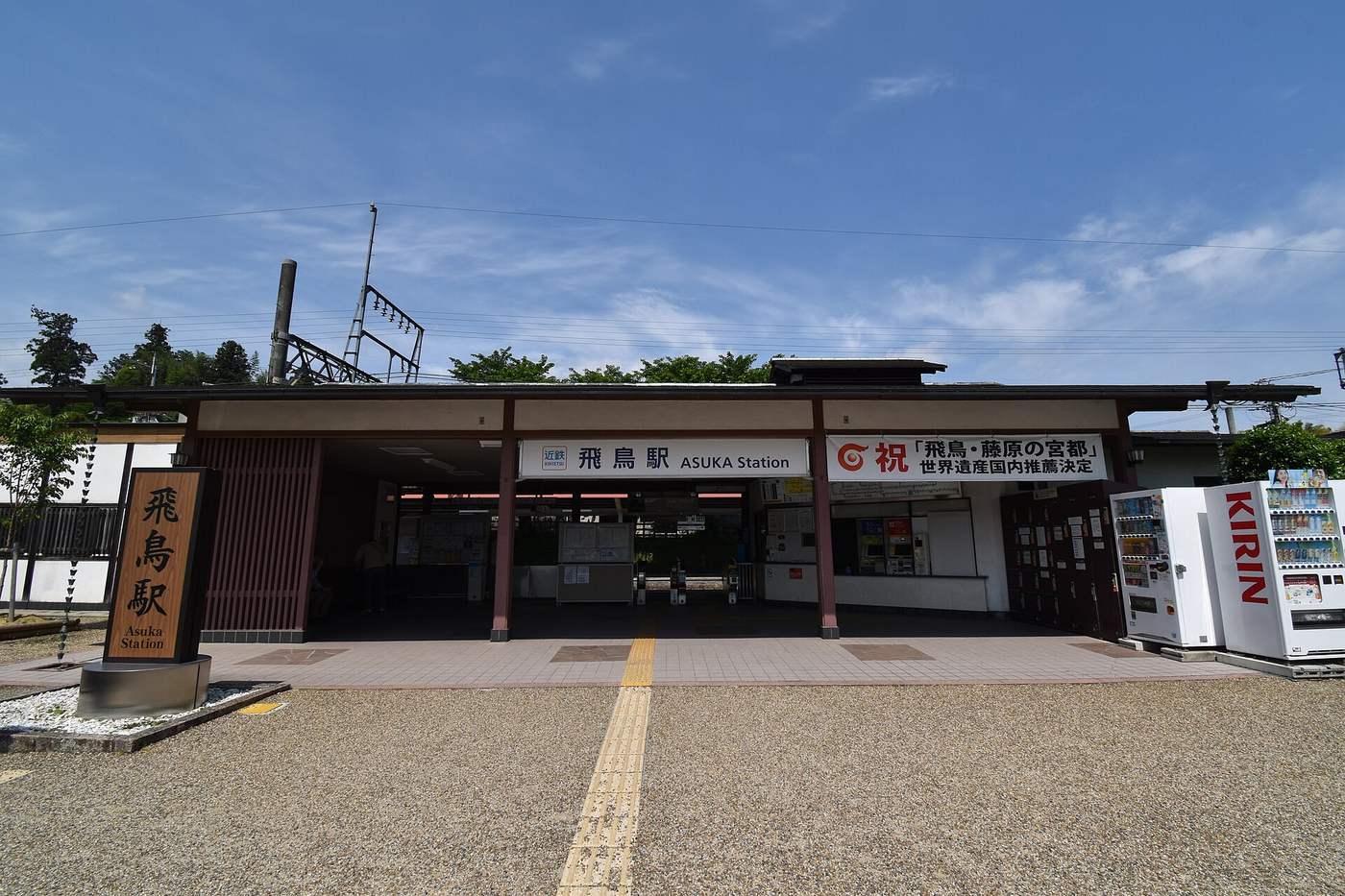 The front of Kintetsu Asuka Station: a low building with dark timber posts under tiled eaves, a white sign reading Kintetsu Asuka Station in Japanese and English, a white banner to its right celebrating the domestic World Heritage nomination, ticket machines and gates visible inside, two drink vending machines at the right and a gravel forecourt with yellow tactile paving