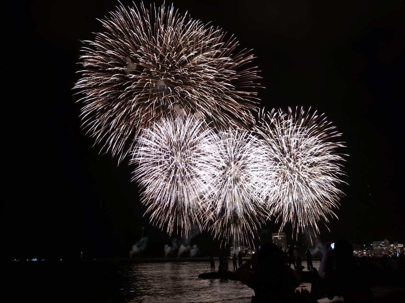 夏夜的熱海海上花火:夜空中同時炸開五顆白金色的大型花火,下方海面與堤防冒著白煙,遠處是亮燈的飯店大樓,前景是舉著手機拍攝的觀眾背影剪影