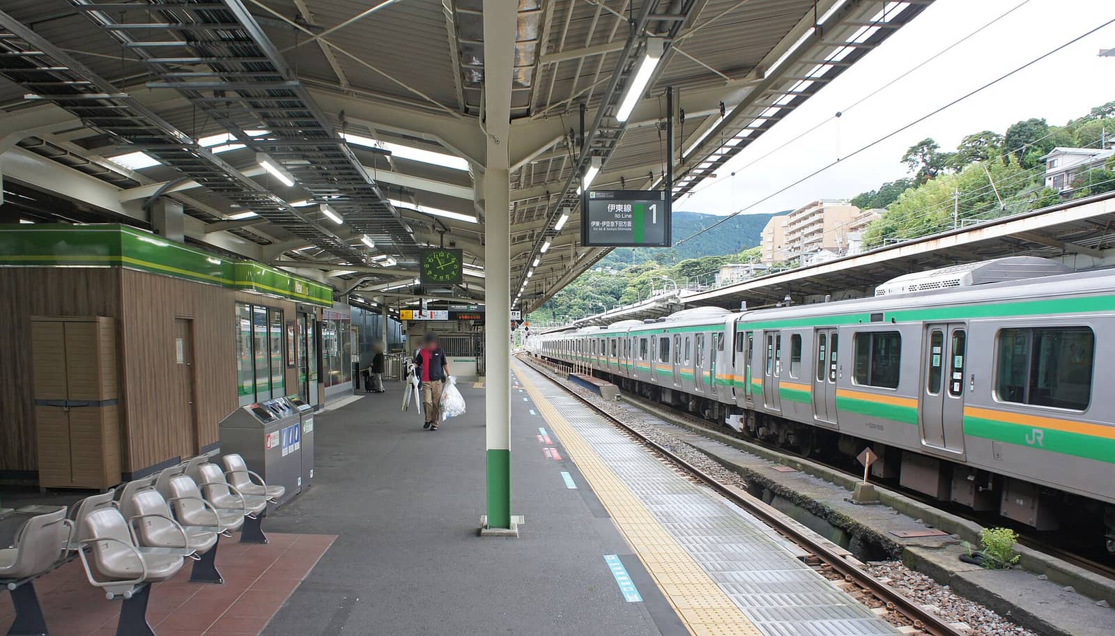 A conventional-line platform at Atami Station, an overhead display reading Ito Line 1 for Ito and Izukyu-Shimoda, a JR train in green and orange Shonan livery standing on the track to the right, a green timber-clad station kiosk and a row of grey seats on the left, and wooded hillside and apartment blocks beyond