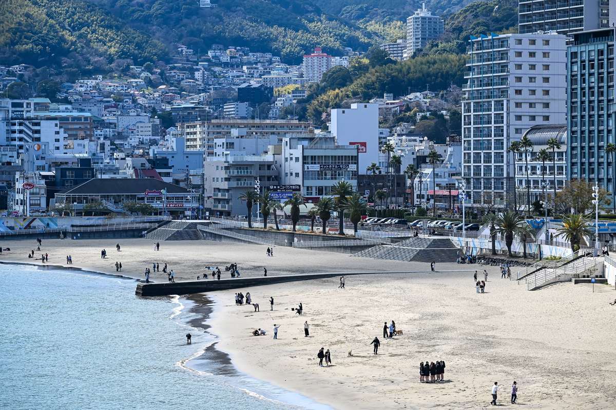 白天從東北側高處看熱海陽光海灘:弧形沙灘上零星有散步和遛狗的人,沙灘中間一道低矮的堤防,後方是棕櫚樹、飯店大樓與蓋滿房子的山坡