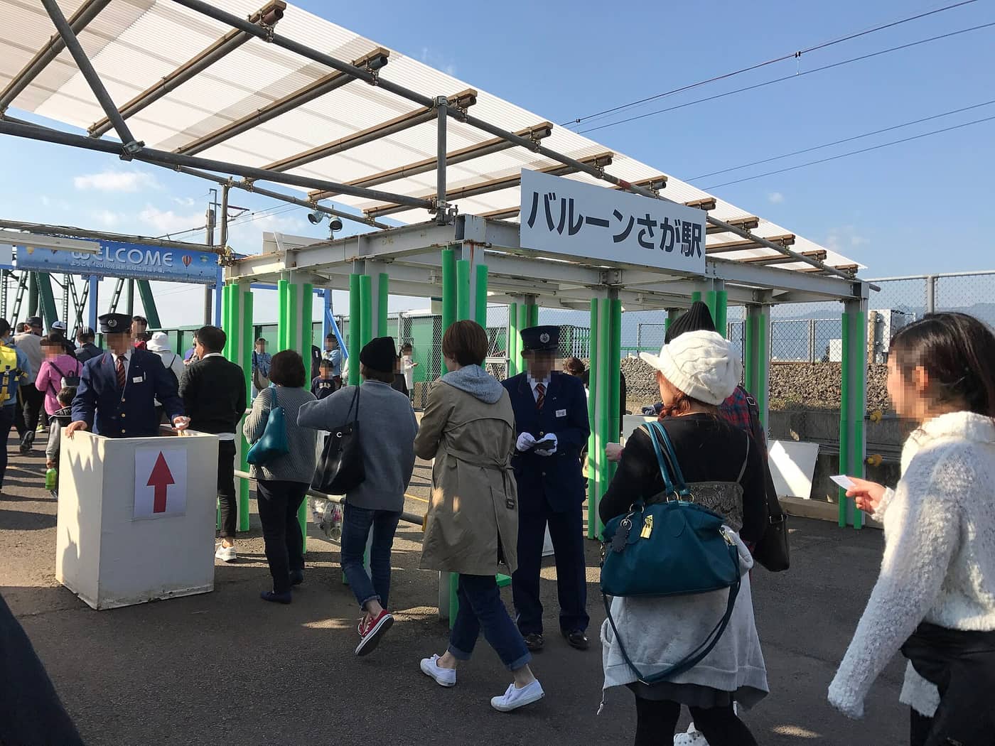 The entrance to Balloon-Saga Station: a white sign reading バルーンさが駅 hanging under a temporary canopy of white scaffold tube and corrugated plastic, a line of fluorescent green scaffold barriers marking out the walkway, two JR staff in dark navy uniforms and peaked caps checking tickets by hand, a blue WELCOME banner to the left, and crowds walking through