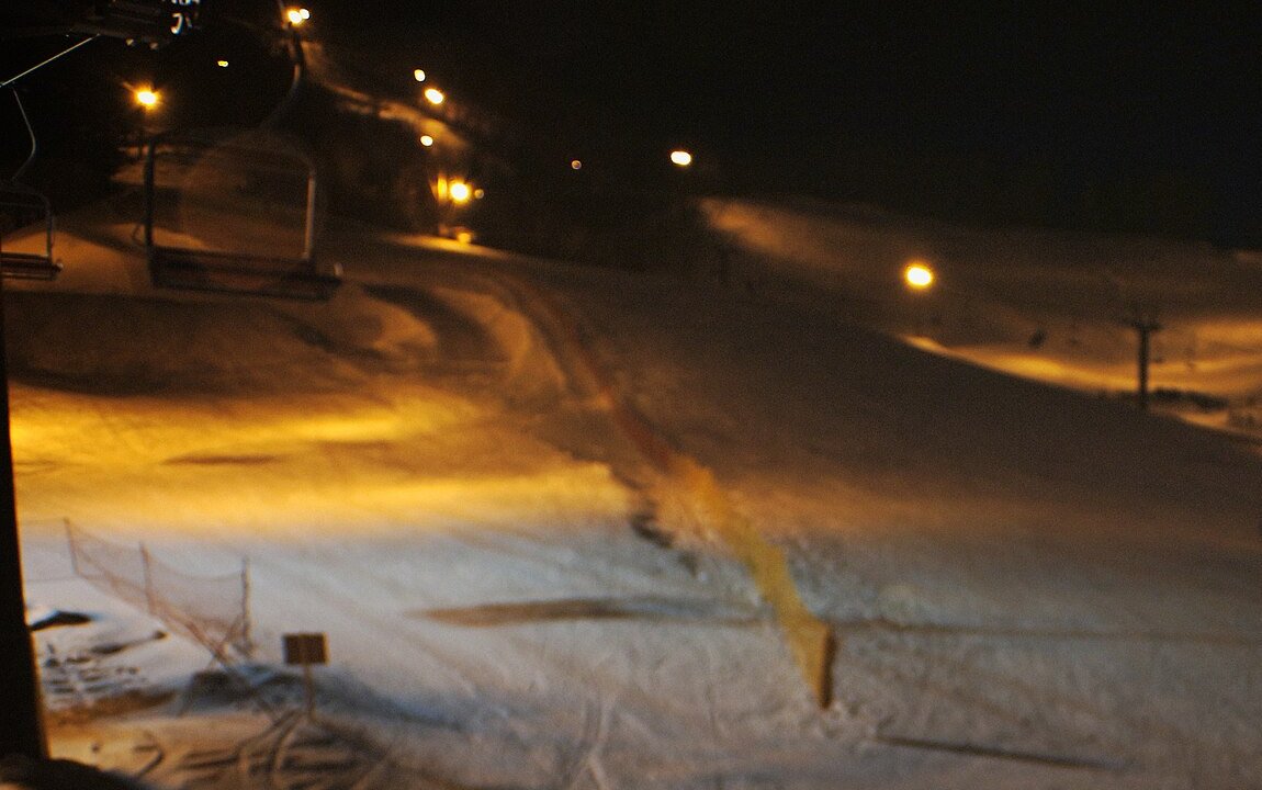 晚上從吊椅纜車上往下看盤溪的雪道:天空全黑,路燈沿著雪道兩側打出橘黃色的光,左側是纜車支柱與空的吊椅,雪面上有壓雪痕與隔網
