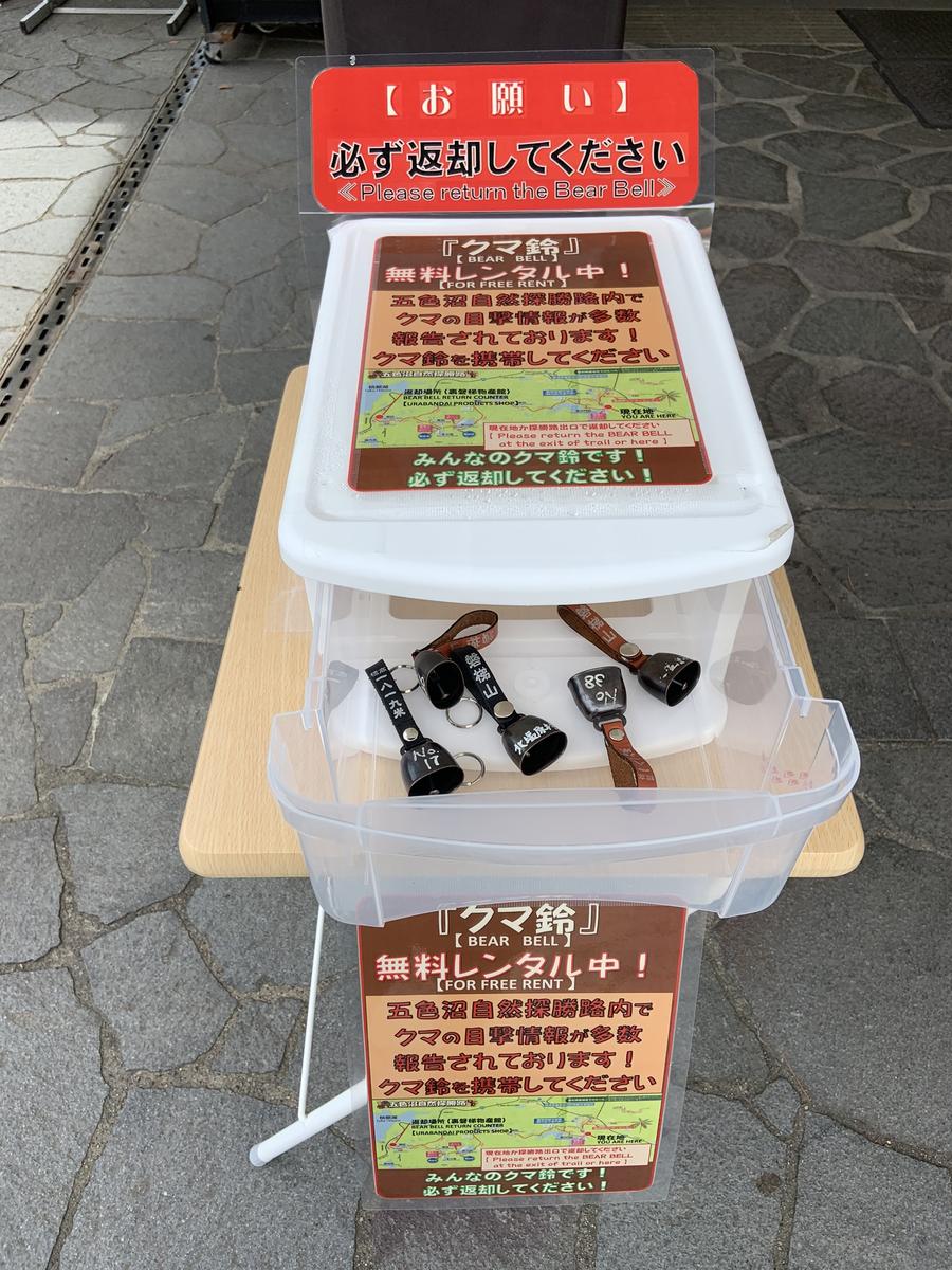 A free bear bell loan station on paving at a trail entrance: a clear plastic drawer unit on a wooden table holding four numbered black bells on leather straps, with Japanese posters offering the bells for free rent and a red sign above reading Please return the Bear Bell