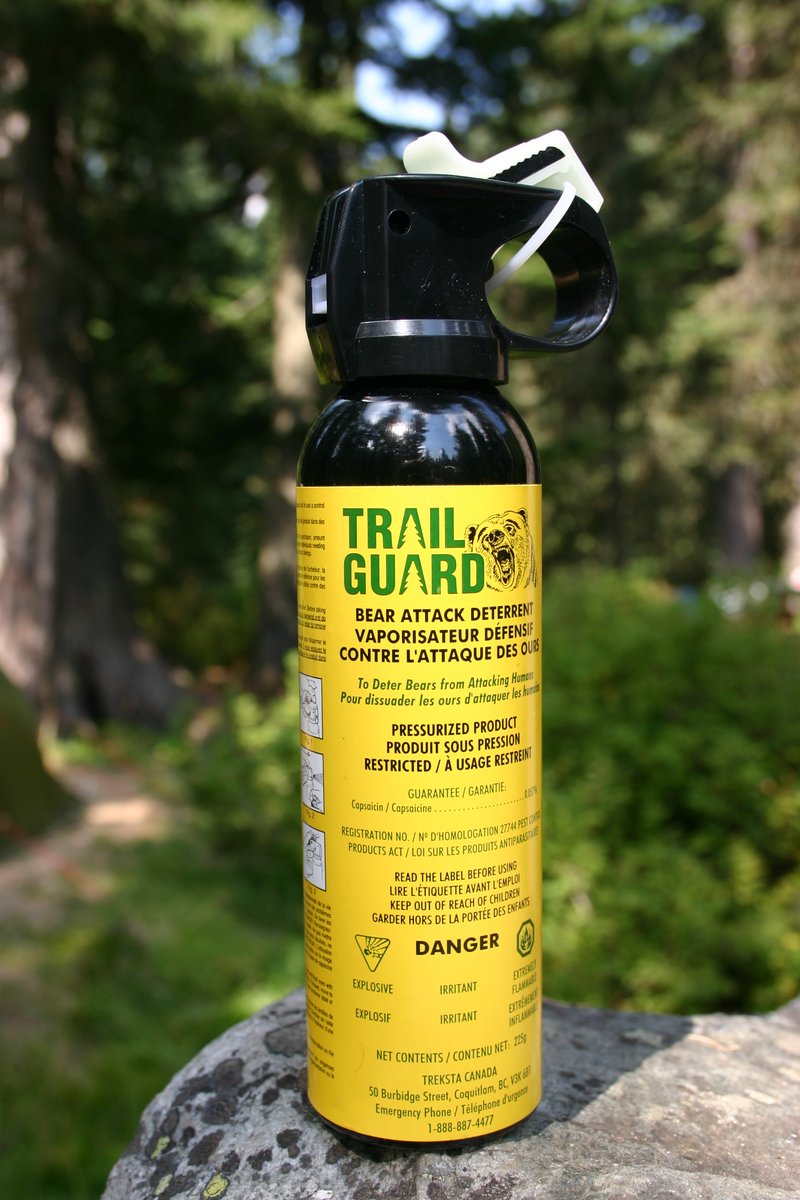 A yellow-labelled bear spray canister standing on a rock in woodland, its black trigger head fitted with a white safety clip, the label reading BEAR ATTACK DETERRENT and PRESSURIZED PRODUCT in English and French above a DANGER panel with explosive, irritant and extremely flammable symbols