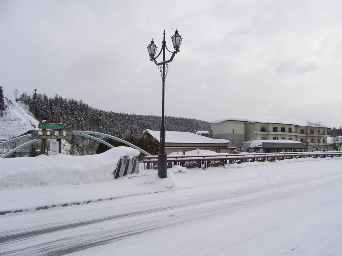 三月初的白金溫泉入口：整條車道被壓實的雪蓋住，橋邊雪堆比欄杆還高，左邊有寫著「白金温泉 SHIROGANE SPA」的木牌和藍綠色拱橋，右後方是屋頂積雪的旅館