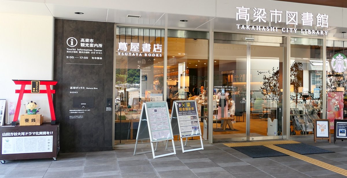 高梁市圖書館一樓入口:左邊深灰色牆上掛著「i 高梁市観光案内所 Tourist Information Center 9:00—17:00 年中無休」的牌子,旁邊是一座小紅鳥居和吉祥物人偶,中間是蔦屋書店的玻璃門與兩塊活動立牌,右上方是「高梁市図書館 TAKAHASHI CITY LIBRARY」的白色字