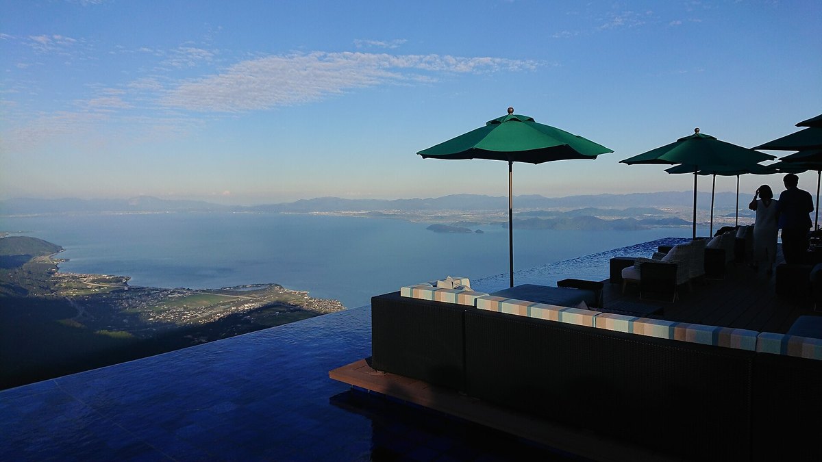 2018 年 10 月傍晚的琵琶湖 Terrace：木棧平台上有綠色陽傘與沙發座，平台外緣是一片貼齊邊緣的水池，遠方是琵琶湖與對岸的山，右側兩位遊客是逆光的剪影