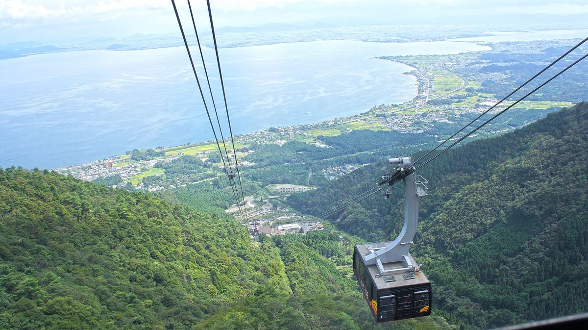2011 年 8 月從琵琶湖 Valley 纜車車廂往下看：鋼索直通山谷裡山麓站一帶的建築，右下是另一台對向的車廂，遠方是琵琶湖與湖岸的田地、聚落，山坡是夏天的綠色