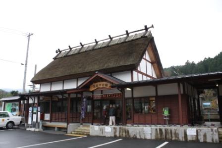 JR 豊後中村駅站房:茅草屋頂、紅褐色木牆的平房,入口上方掛著木製站名牌,站前是停車場