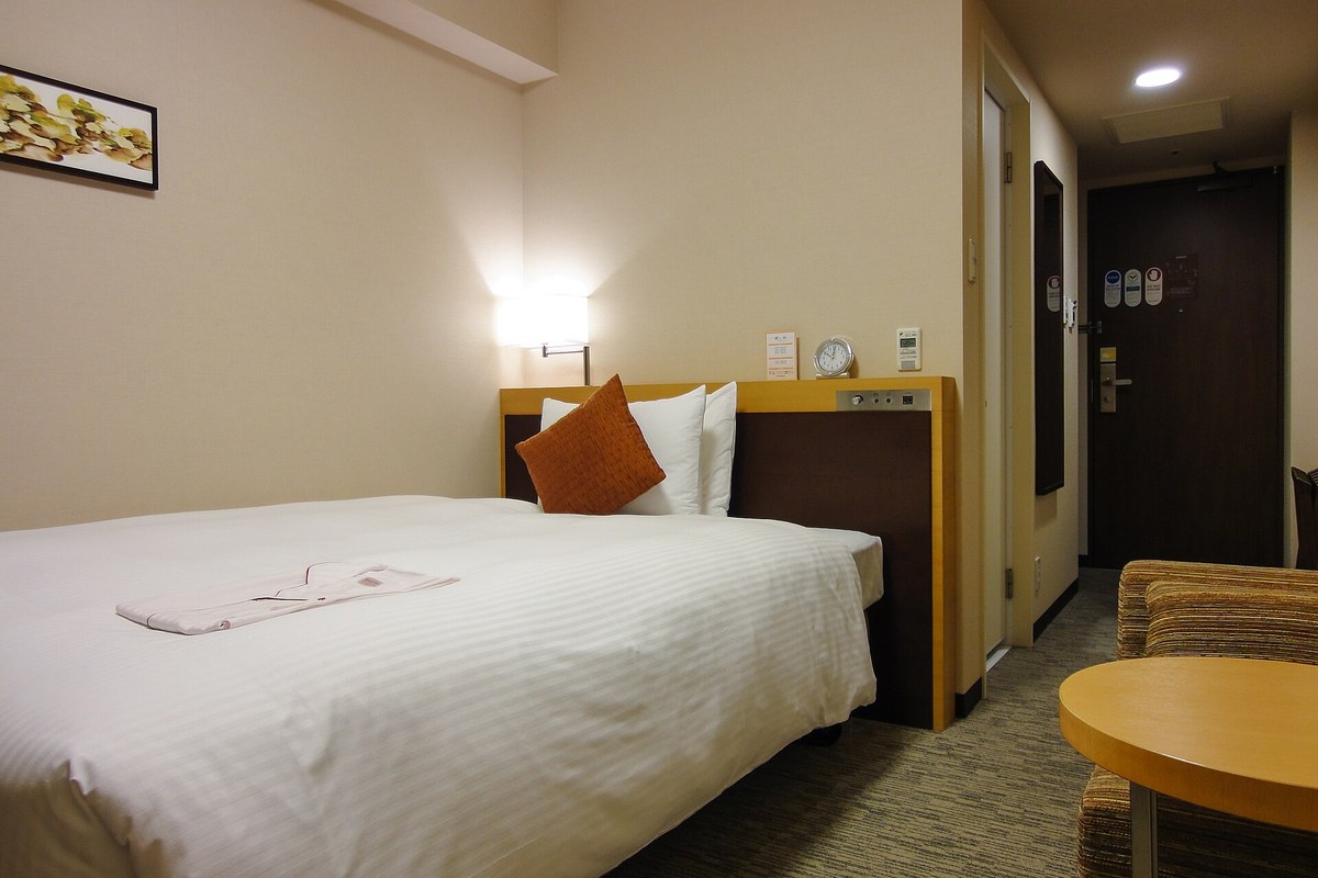 A Japanese business hotel single room: the bed takes up most of the floor, the headboard panel holds the light switches and a clock, a narrow walkway on the right leads past the white unit bath door to the room door, and the corner fits only a small round table and one chair