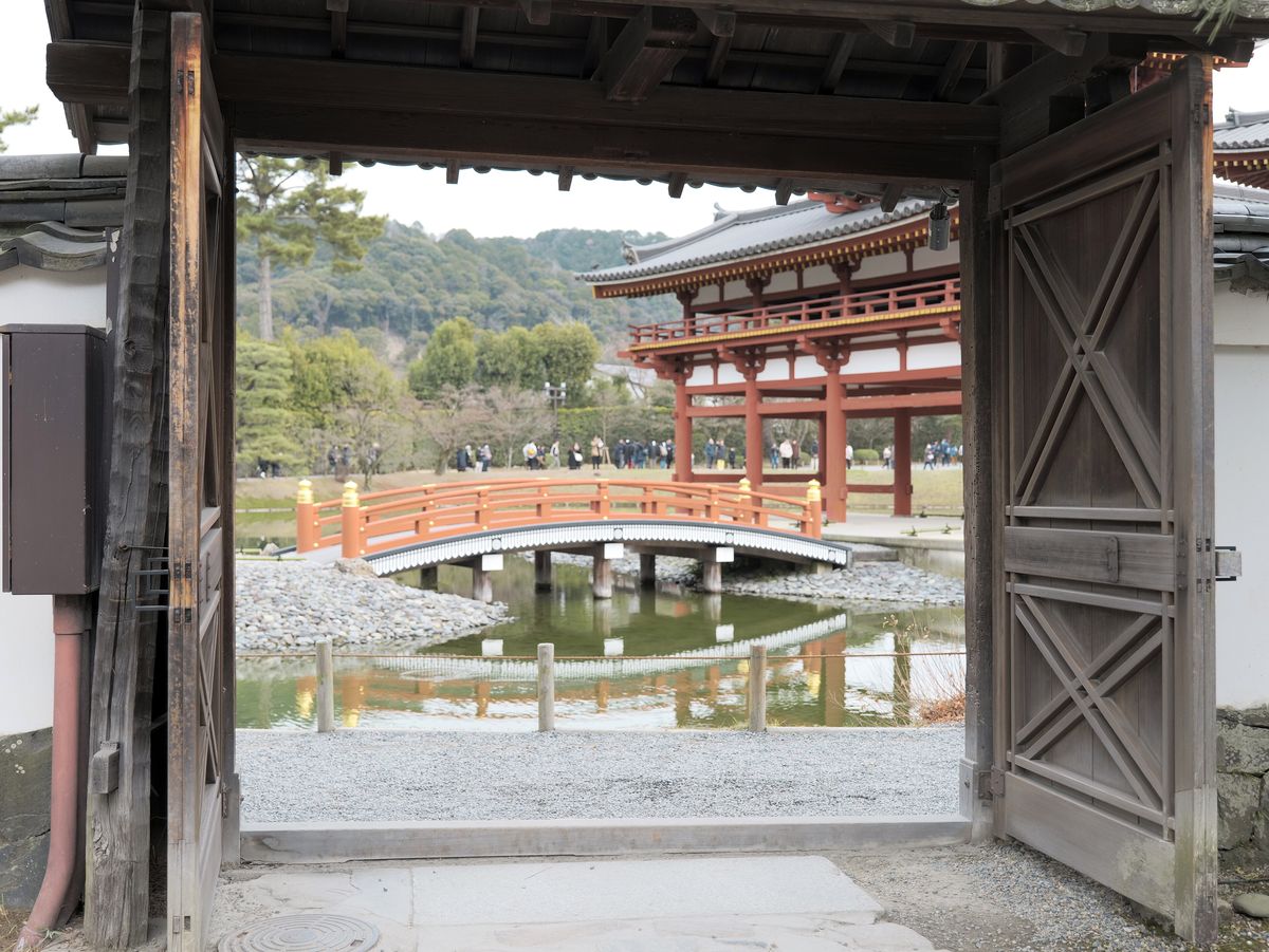 從平等院庭園一道木門往外看:門框裡是朱紅色的拱橋與阿字池,右側是鳳凰堂翼廊,遠處草地上有一排小小的遊客身影,背後是綠色山丘,陰天