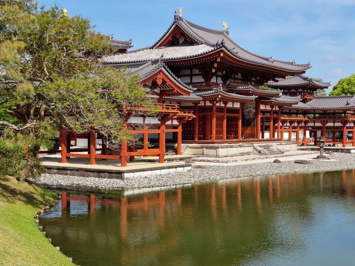 從阿字池岸邊斜看平等院鳳凰堂:中堂正面的朱紅柱子與門扉、右側翼廊,前景一棵松樹,池面映出柱子的倒影,晴天