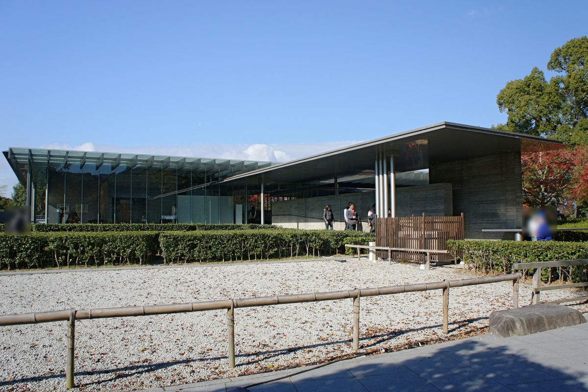 平等院博物館鳳翔館外觀:低矮的玻璃與清水混凝土建築,大片斜屋簷,前面是白色碎石與竹籬,右側有紅葉,幾位遊客在入口附近(臉部已模糊)
