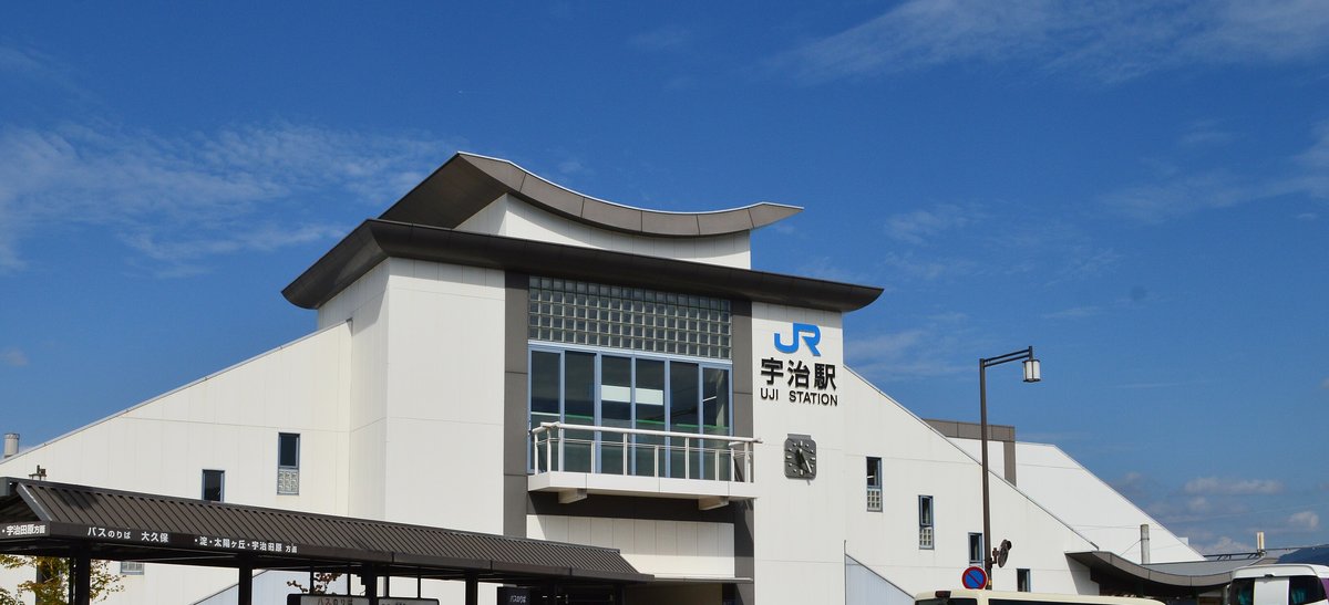 JR 宇治站站房:白色建築,屋頂是向上翹起的弧形,正面有藍色 JR 標誌與「宇治駅 UJI STATION」站名,左下角是站前公車站的屋簷,晴天