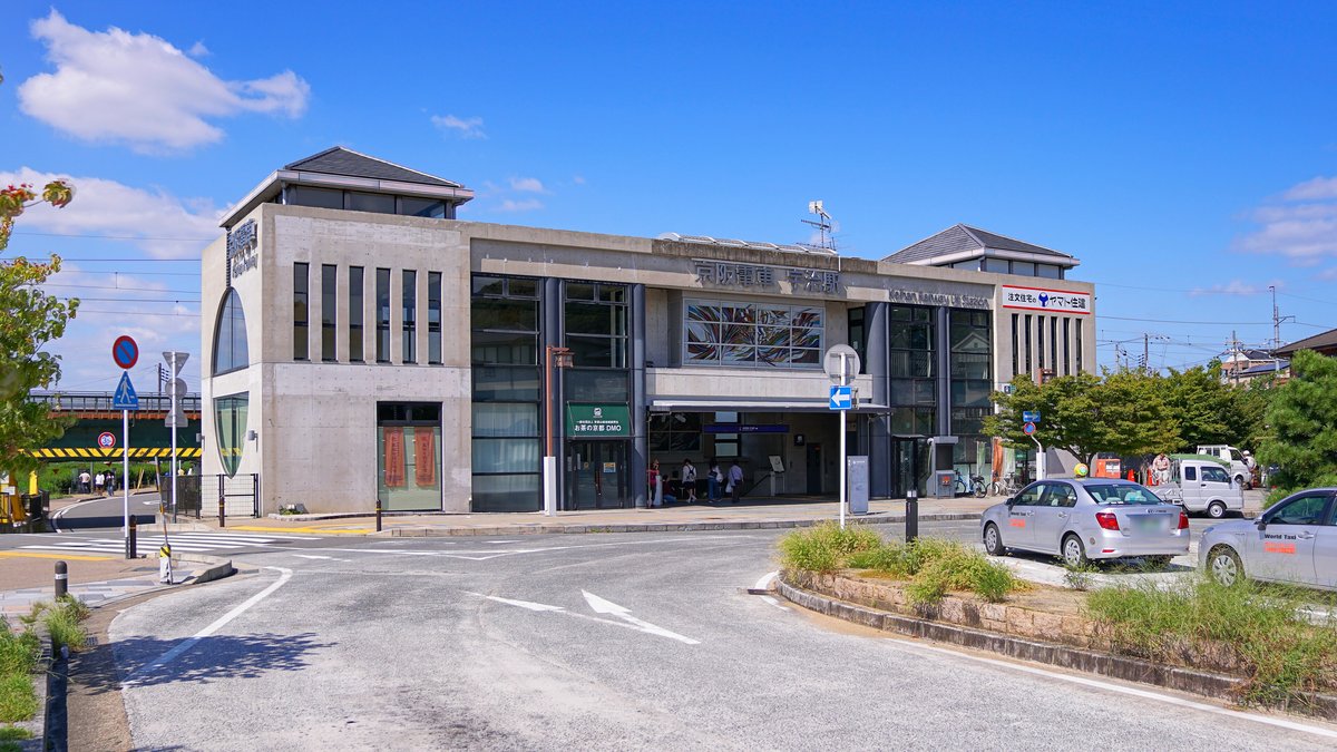京阪宇治站站房:灰色清水混凝土建築,正面刻著「京阪電車 宇治駅」,左側有大片圓弧形玻璃窗,站前圓環停著兩台計程車,晴天