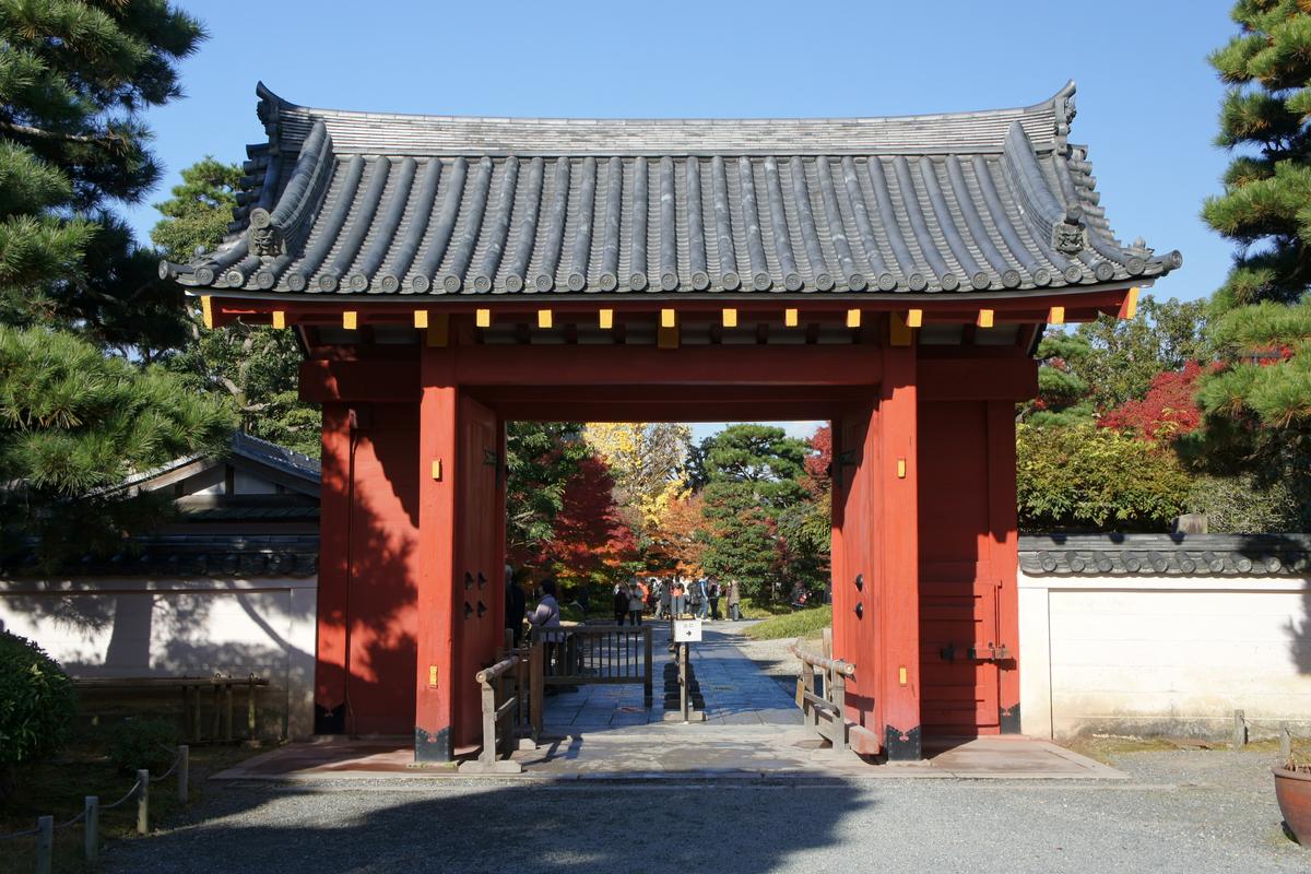 平等院表門:灰瓦屋頂、朱紅柱子的四腳門,門扇向內打開,門內石板路上立著「出口」箭頭指示牌,遠處有紅葉與幾位遊客,兩側是白色土牆