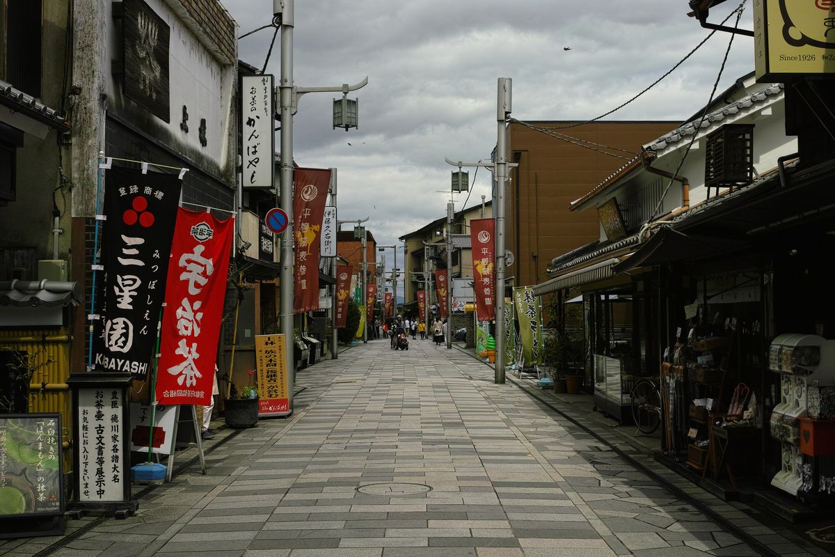 平等院表參道:石板鋪成的步行街,兩側是茶舖,掛著「三星園」「宇治茶」與印有「平等院表参道」字樣的紅色旗幟,街上只有遠處幾位行人,陰天