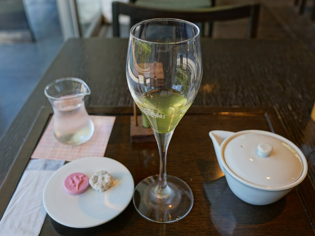 茶房藤花的桌上:高腳酒杯裝著淡綠色的冷泡玉露,旁邊一只白色急須與一小瓶水,小碟上放著粉紅色與白色兩顆和菓子