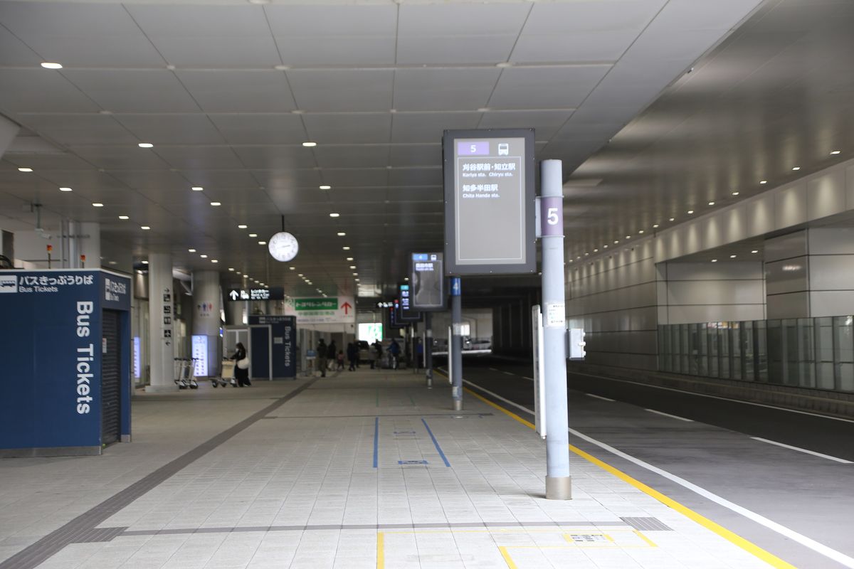 Access Plaza 1 樓的巴士乘車處:有頂棚的長廊一側是車道,前方灰色柱子上是 5 號站牌,看板寫「刈谷駅前・知立駅」「知多半田駅」,往後還有 4 號等站牌;左邊深藍色亭子寫「バスきっぷうりば Bus Tickets」,遠處是租車指標