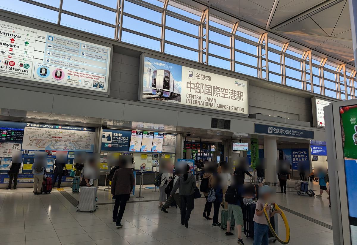 名鐵中部國際機場站入口:上方大看板寫「名鉄線 中部国際空港駅」,左邊一整排售票機上方是運賃表與「名鉄線きっぷうりば」「特別車両券(ミューチケット)うりば」,中間是有人窗口,右邊藍色看板寫「名鉄線のりば」與「ミュースカイは1番線から発車します」,大廳裡有拉行李的旅客