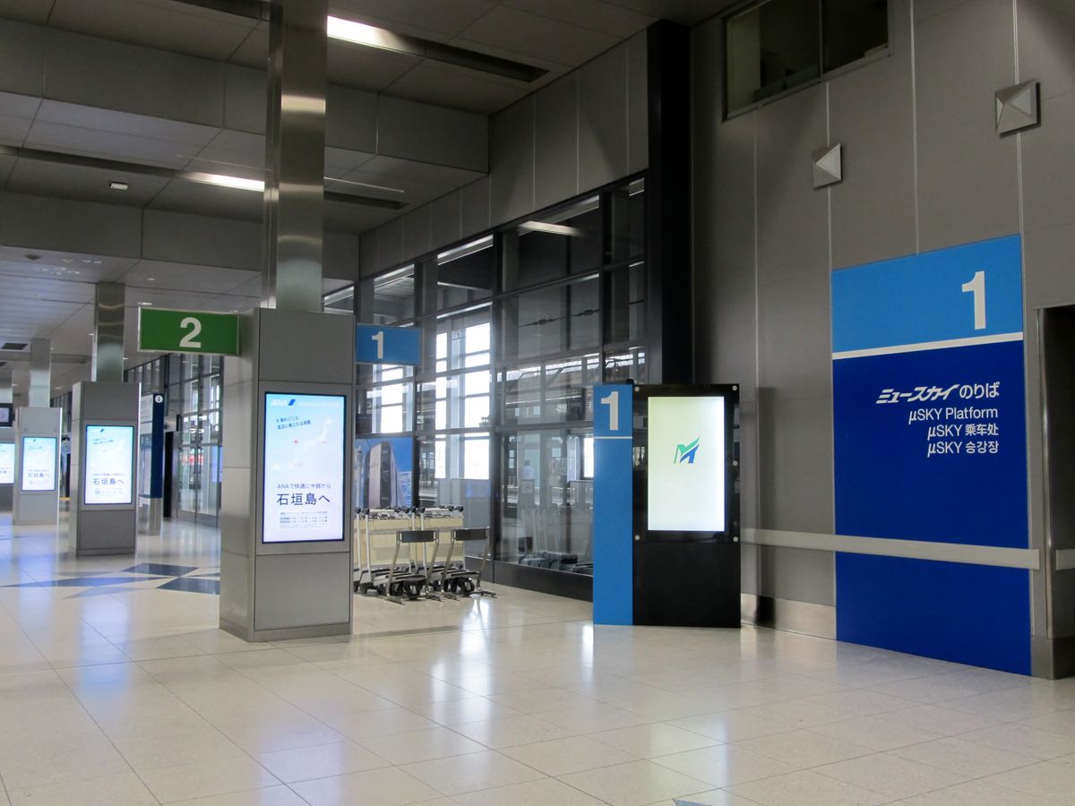 名鐵中部國際機場站月台層:右邊藍色大看板寫「1 ミュースカイのりば μSKY Platform μSKY 乘车处 μSKY 승강장」,中間有藍色的 1 號牌與綠色的 2 號牌,柱子上的螢幕在播廣告,月台旁放著一排行李推車