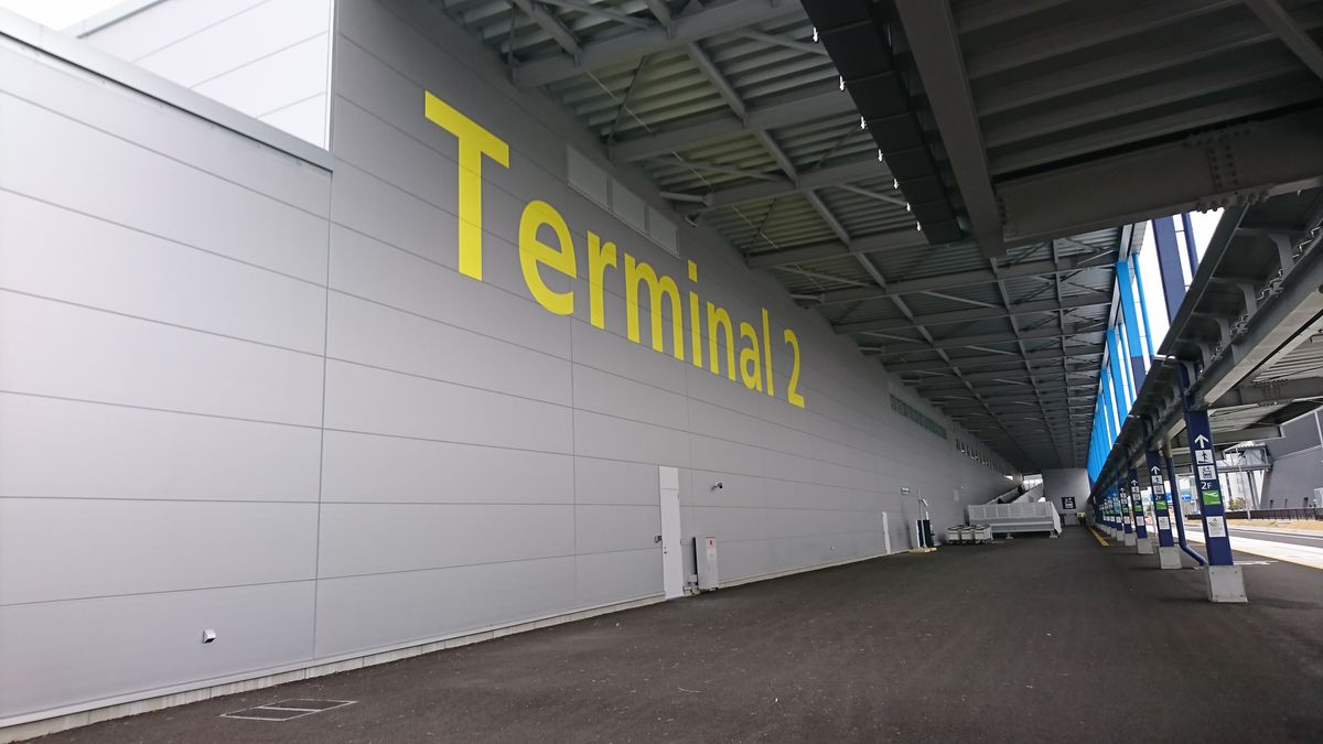 中部機場第 2 航廈的外牆:灰色鋼板牆上漆著大大的黃色「Terminal 2」,右邊是有頂棚的人行道與藍色柱子,柱上指標寫著往 2 樓出發大廳