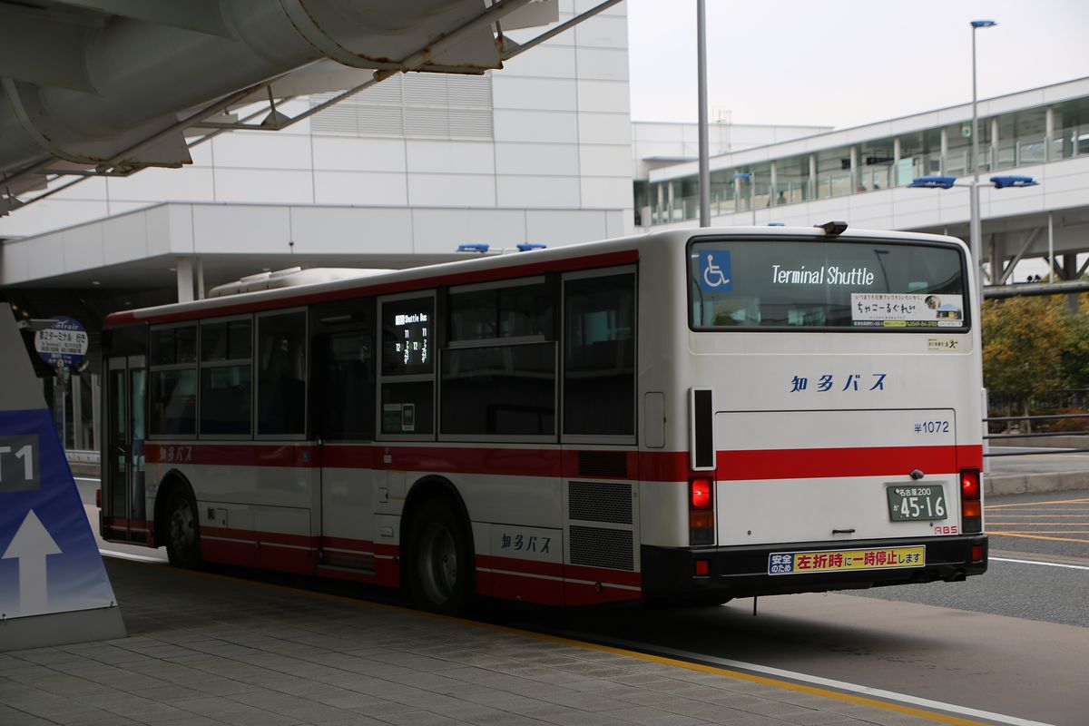 停在第 1 航廈 Access Plaza 站牌旁的航廈接駁車:紅白相間的知多巴士,車尾電子看板寫「Terminal Shuttle」,側面看板顯示 T1、T2;左邊站牌是寫著「第2ターミナル 行き」的圓形牌子,前方藍色立牌印著「T1」