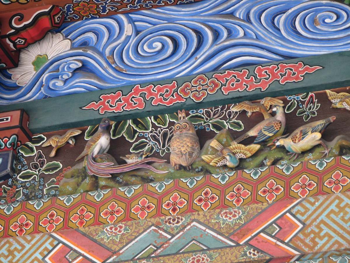 秩父神社社殿的彩色木雕近景,中央是一隻身體朝前、頭轉向正面的貓頭鷹站在綠色苔石上,左右各有數隻彩繪小鳥與長尾鳥;上方是藍白相間的波浪雕刻,下方是金底六角龜甲紋與花草彩繪