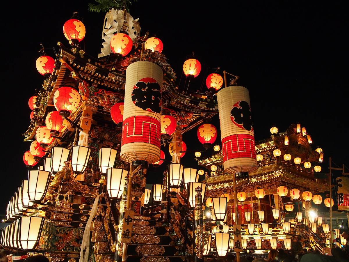 兩座掛滿提灯的秩父夜祭笠鉾在夜裡並排,前方那座掛著寫「中近」的木牌,車頂旁立著兩支白底紅帶的大型圓筒提灯,下層排滿方形白提灯,車體是彩色木雕加金箔,背景是全黑的夜空