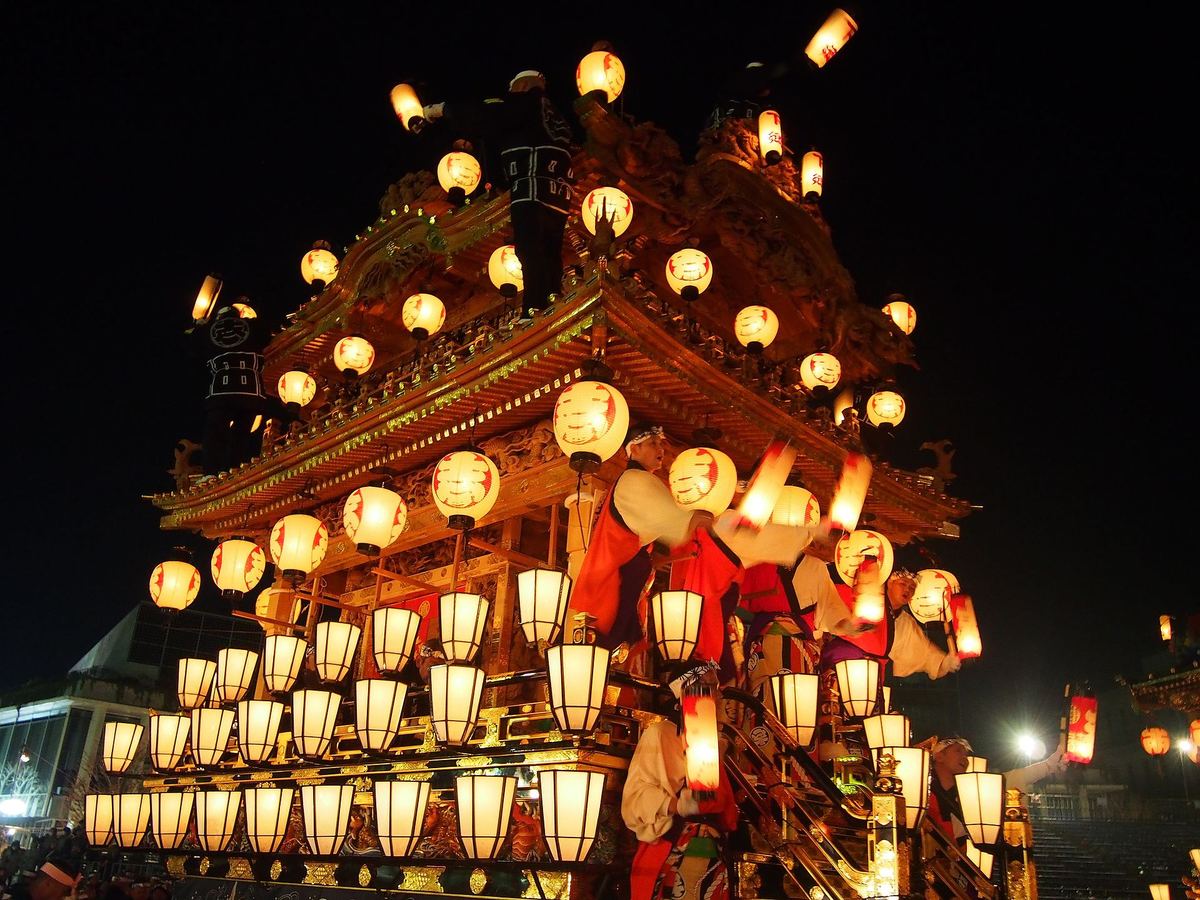 秩父夜祭的屋台在夜裡由下往上仰拍,兩層屋簷與整面欄杆掛滿數十盞亮著的紅色圓提灯與白色方提灯,屋頂上站著兩名穿黑色半纏的男子,欄杆邊幾個穿紅白裝束的人正把手提提灯往外揮