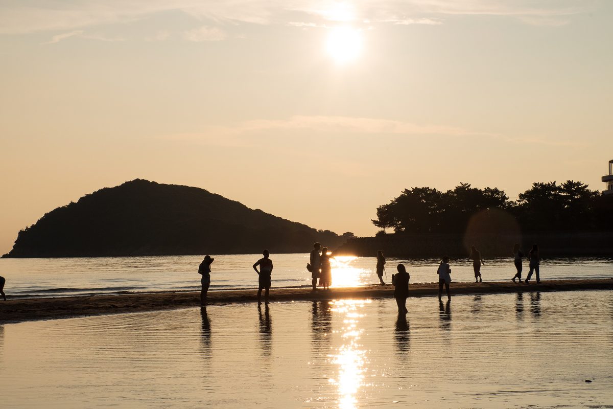 父母ヶ浜的夕陽：退潮後的沙灘上留著一層薄水，太陽在淡橘色的天空偏上方，水面映出一道金色倒影，十幾個人的剪影站在沙洲上拍照，左邊是一座黑色的小島
