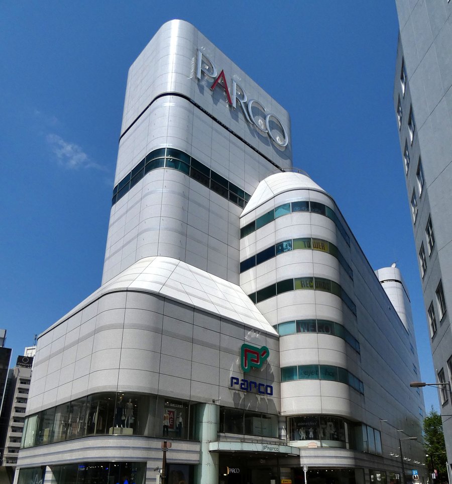P'PARCO 的外觀:銀白色的高塔頂端掛著大大的 PARCO 字樣,低樓層轉角有紅綠色的 P' 標誌與藍色 parco 字,右側圓弧外牆的窗帶上有 TOWER RECORDS 字樣