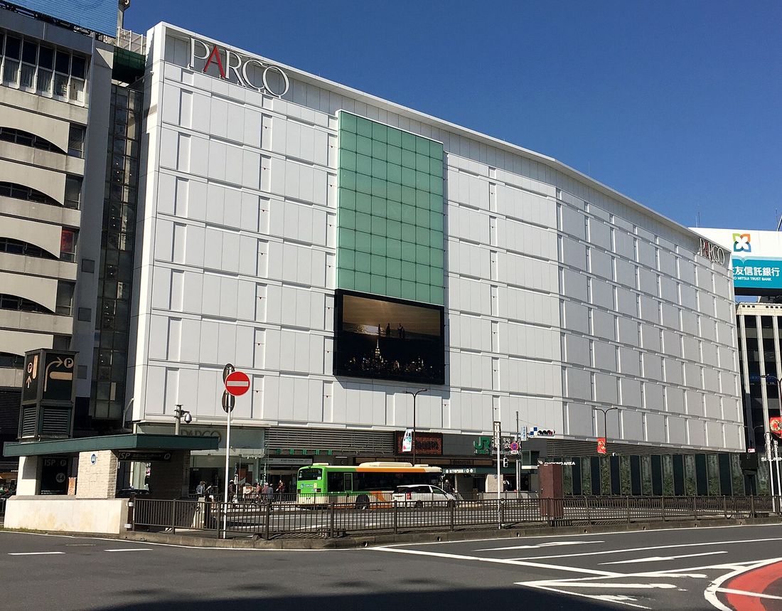 晴天下的池袋 PARCO 本館：白色方格外牆，左上角紅色 PARCO 字樣，中央一片綠色玻璃帷幕與大螢幕，一樓掛著綠色 JR 池袋駅站名牌，前面是站前道路與一台綠色公車